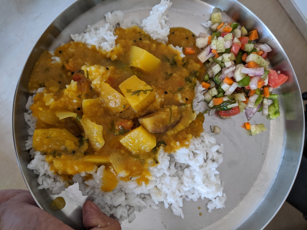 A plate of rice on which there is a sambar of potatoes, onion and brinjal along with a green cut salad